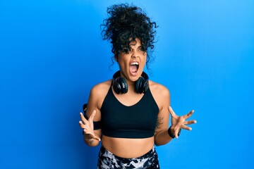 Fototapeta na wymiar Young hispanic woman wearing gym clothes and using headphones crazy and mad shouting and yelling with aggressive expression and arms raised. frustration concept.