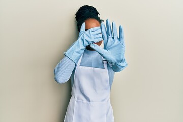 African american woman with braided hair wearing cleaner apron and gloves covering eyes with hands...