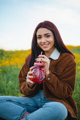 Smiling teenager girl drinking water in a red jar sitting on the grass of a hill. She is looking at camera.