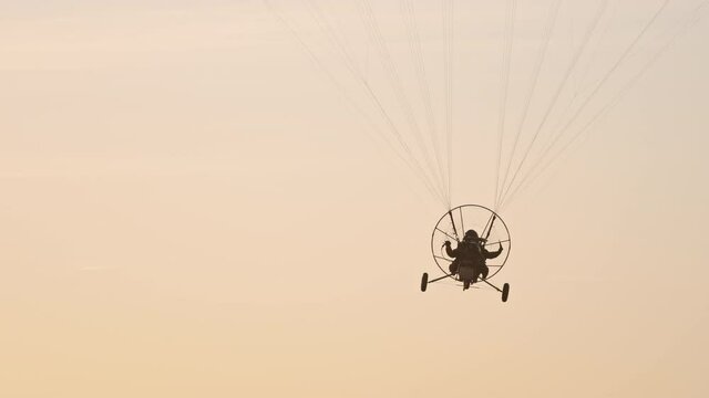 Close-up of paramotor trike hanged under parachute flying away from camera. Tandem motor powered paragliding at twilight. 