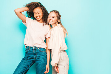 Two young beautiful smiling international hipster female in trendy summer clothes. Sexy carefree women posing near blue wall in studio. Positive models having fun. Concept of friendship