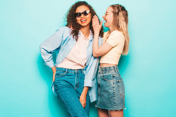 Two young beautiful smiling international hipster female in trendy summer clothes. Sexy carefree women posing near blue wall in studio. Positive models having fun. Tells her friend secret