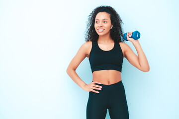 Fototapeta na wymiar Fitness smiling black woman in sports clothing with afro curls hairstyle.She wearing sportswear. Young beautiful model with perfect tanned body.Female holding dumbbells in studio near light blue wall