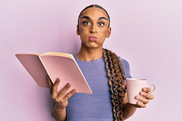 Hispanic man wearing make up and long hair reading book drinking cup of coffee puffing cheeks with...