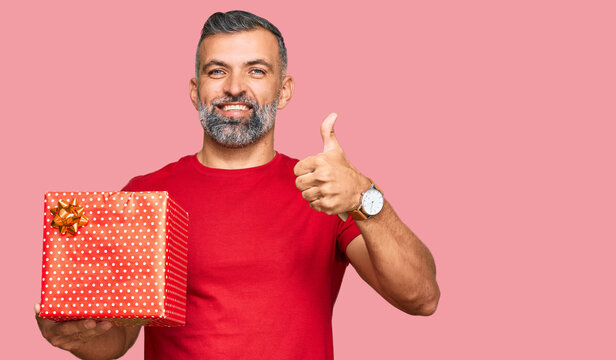 Middle age handsome man holding gift smiling happy and positive, thumb up doing excellent and approval sign