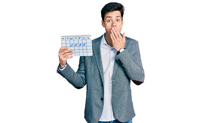 Young hispanic business man holding travel calendar covering mouth with hand, shocked and afraid for mistake. surprised expression