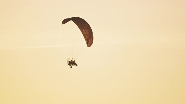 Silhouette of paramotor flying in air. Parachute against orange sunset sky. Tandem motor powered paragliding at twilight. 