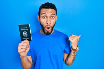 Hispanic man with beard holding italy passport surprised pointing with hand finger to the side, open mouth amazed expression.