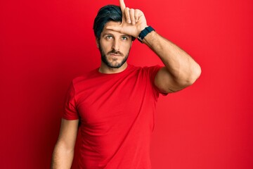 Fototapeta premium Young hispanic man wearing casual red t shirt making fun of people with fingers on forehead doing loser gesture mocking and insulting.