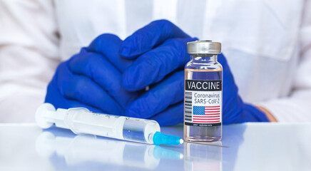 Coronavirus vaccine with USA flag on label, American vaccination concept, doctor hand in blue gloves and glass table with medical syringe