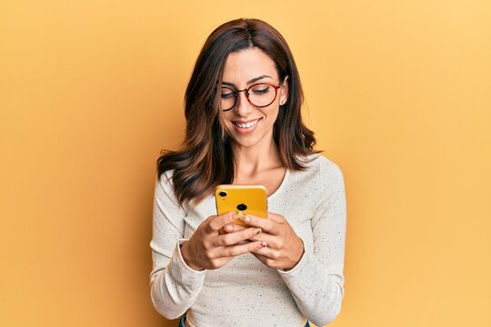 Young brunette woman using smartphone over yellow background smiling with a happy and cool smile on face. showing teeth.