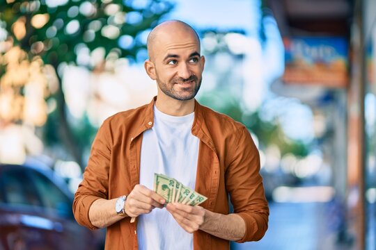 Young hispanic bald man smiling happy counting usa dollars at the city.