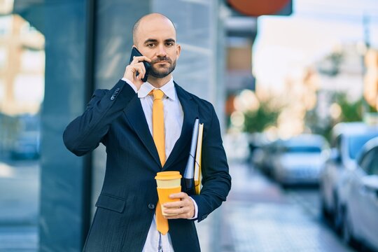 Young hispanic bald businessman with serious expression talking on the smartphone drinking coffee at the city