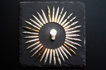 steel pen nibs arranged in a circle, with a small light bulb  in the middle, isolated on black stone texture