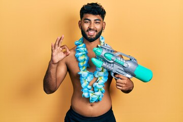 Arab man with beard wearing swimsuit and hawaiian lei holding watergun doing ok sign with fingers,...
