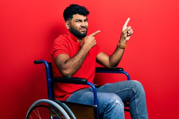 Arab man with beard sitting on wheelchair pointing aside worried and nervous with both hands, concerned and surprised expression