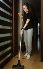 Teenage Girl With Wireless Headphones  Taking Break Of Vacuuming And Checking Her Smartphone.