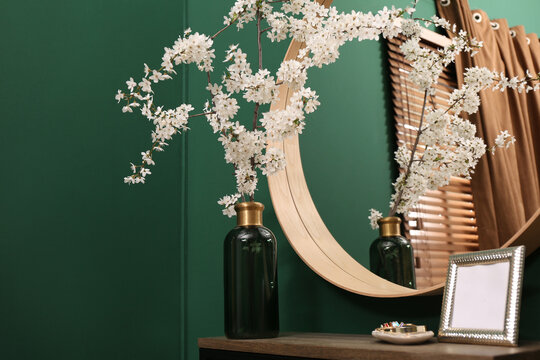 Blossoming tree twigs in vase and empty photo frame on wooden table near mirror indoors