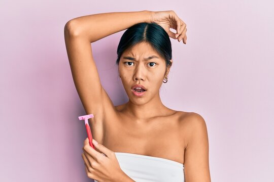 Young chinese woman wearing shower towel using razor in shock face, looking skeptical and sarcastic, surprised with open mouth
