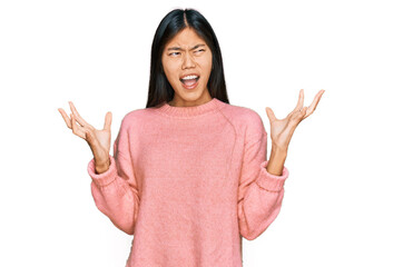 Beautiful young asian woman wearing casual winter sweater crazy and mad shouting and yelling with aggressive expression and arms raised. frustration concept.