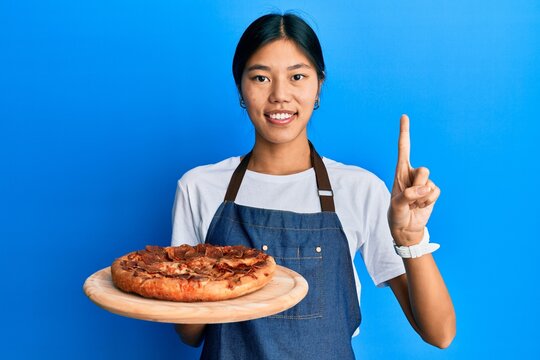 Young chinese woman wearing waiter apron holding italian pizza smiling with an idea or question pointing finger with happy face, number one