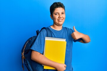 Teenager hispanic boy wearing school bag and holding books smiling happy and positive, thumb up doing excellent and approval sign