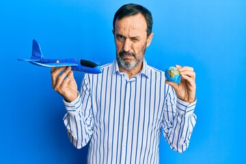 Middle age hispanic man holding plane toy and world ball skeptic and nervous, frowning upset...