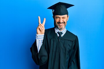 Middle age hispanic man wearing graduation cap and ceremony robe smiling with happy face winking at the camera doing victory sign. number two.