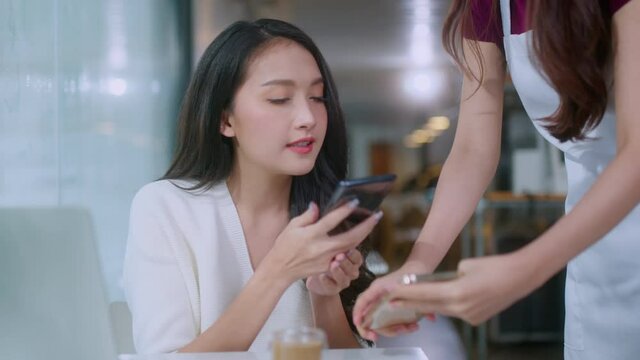 Contactless payment with mobile in restaurant. Waitress standing, wearing protective face mask and apron with asian smiling customer with her smartphone on hand about to pay by qr code payment system