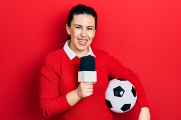 Young hispanic woman holding reporter microphone and soccer ball smiling with a happy and cool...