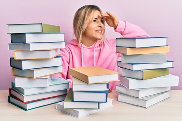 Young caucasian woman sitting on the table with books very happy and smiling looking far away with hand over head. searching concept.