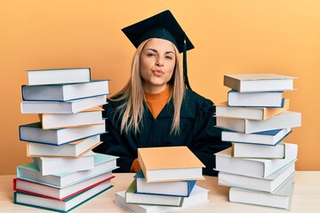 Young caucasian woman wearing graduation ceremony robe sitting on the table looking at the camera blowing a kiss on air being lovely and sexy. love expression.