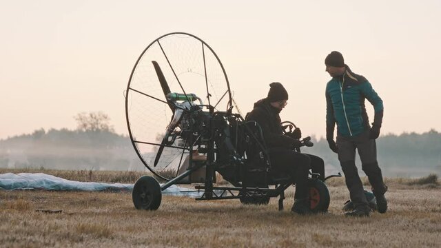 Instructor fastening client's safety belts into seat in trike. Tandem motor powered paragliding at twilight. 