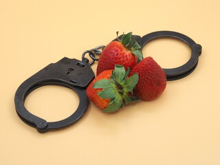 Police metal handcuffs and red ripe strawberries on a yellow table background. The concept of love...