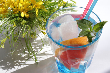 Summer iced grapefruit cocktail with mint leaves and yellow flowers on white background. Refreshing drink with ice cubes. Glass of lemonade with citrus fruit. Hard light, shadow. Top view, copy space	
