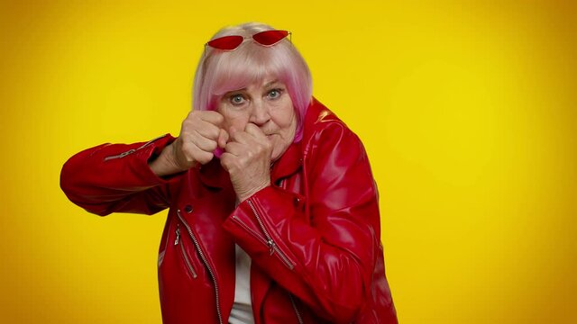 Funny elderly rocker granny woman trying to fight at camera, shaking fist, boxing with expression, punishment. Senior stylish old grandmother isolated on yellow background. People lifestyle emotions