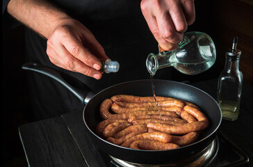The chef adds oil to pan with raw meat sausage. Preparation for cooking sausages in kitchen of a restaurant or cafe on table with vegetables