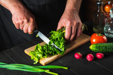 Chef or cook cuts fresh napa cabbage with knife for salad on a vintage kitchen table with fresh vegetables. Cooking and restaurant or cafe concept