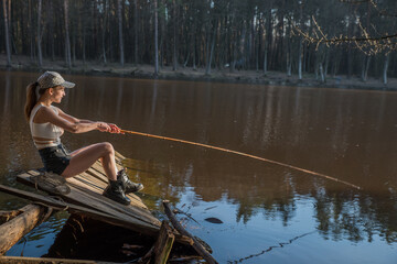 A girl with a fishing rod on a wooden pier is fishing in a forest lake.