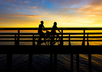 Two young women cycling on bridge at sunset