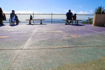 a man in a wheel chair and a man and his son sitting on a bench near the blue ocean with blue sky at Dana Strands Beach in Dana Point California - obrazy, fototapety, plakaty