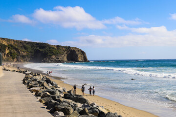 people walking along the beach on silky sand near vast blue ocean water with waves rolling in and blue sky at Dana Strands Beach in Dana Point, California - obrazy, fototapety, plakaty