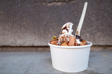 Close - up outdoor view at White Frozen Yogurt serve with topping with melon, kiwi and chocolate in paper white cup, on the floor.