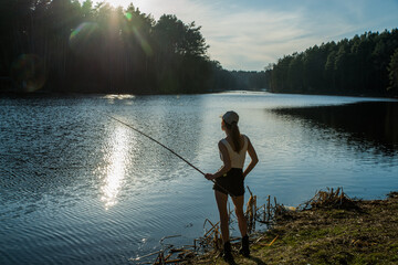 A girl stands on the shore of a forest lake and catches fish with a fishing rod.