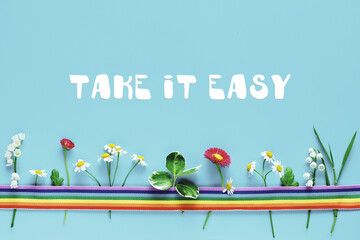 Text Take it Easy on blue paper. Rainbow ribbon, wild flowers. Flat lay, top view, motivation text. Simple naive minimal arrangement.