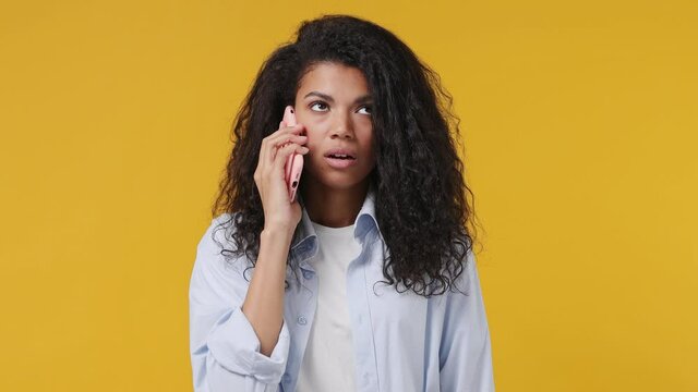 Perplexed angry sad young african woman curly hair 20s years old in blue white t shirt talk on mobile cell phone ask what bad news scream swear isolated on yellow color wall background studio portrait