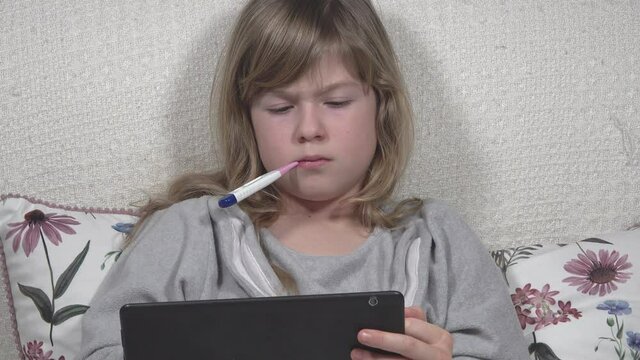 The cute little blonde girl is sick, lies in bed, measures her fever while taking a tablet 