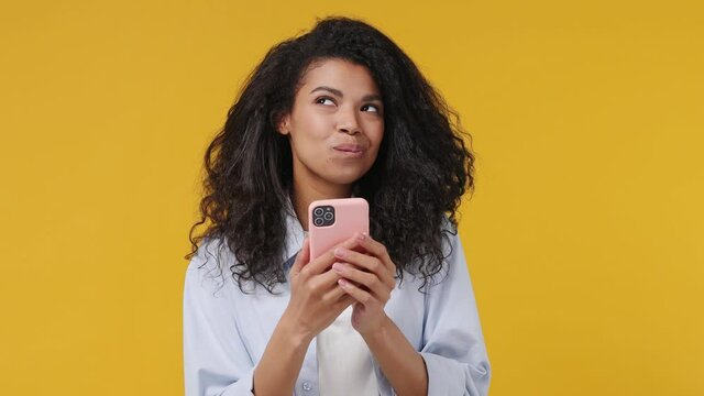 Dreamful pensive african woman curly hair 20s years old wears blue white t shirt hold using mobile cell phone typing browsing chatting send sms isolated on yellow color wall background studio portrait