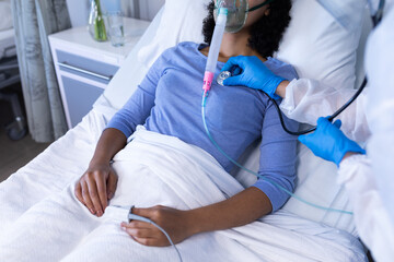 Caucasian doctor examining with stethoscope african american female patient with oxygen ventilator
