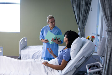 Caucasian female doctor holding file talking with mixed race patient sitting up in hospital bed